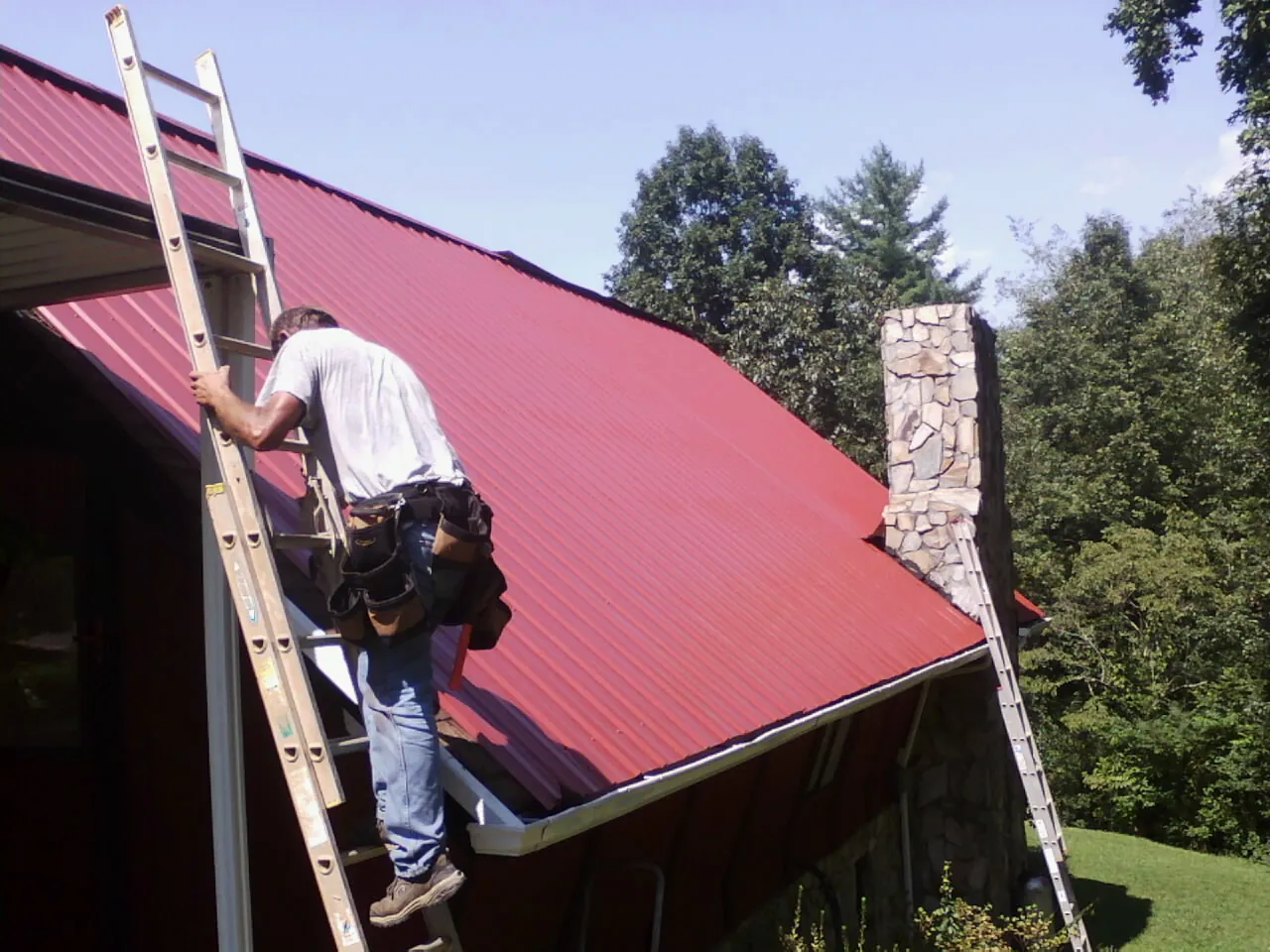 Roofer on a ladder beside a red metal roof and stone chimney — Soffit & Fascia Repair by Roofing Brothers in Marianna